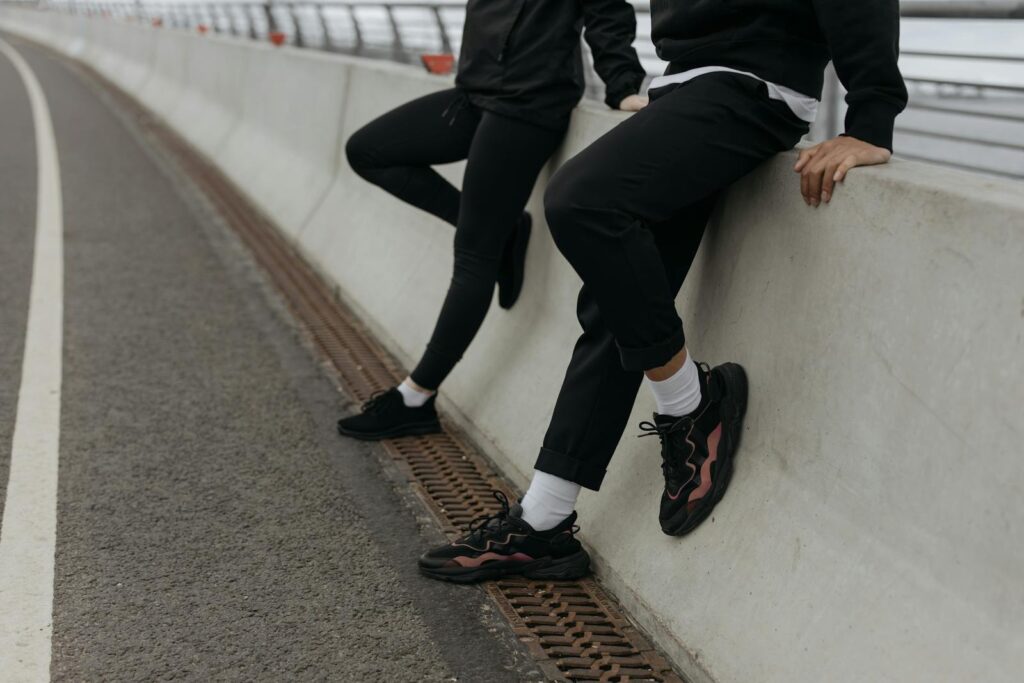 Two people in casual attire resting on a bridge barrier with sneakers and activewear.