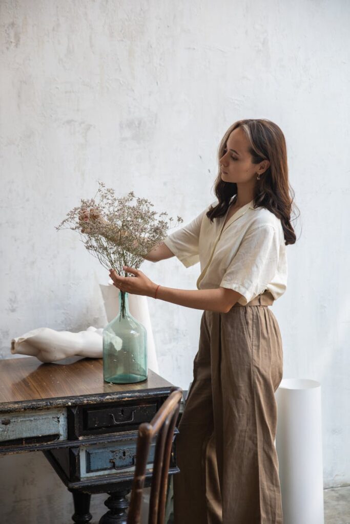 Woman in a linen outfit arranging flowers in a glass vase inside a vintage room.