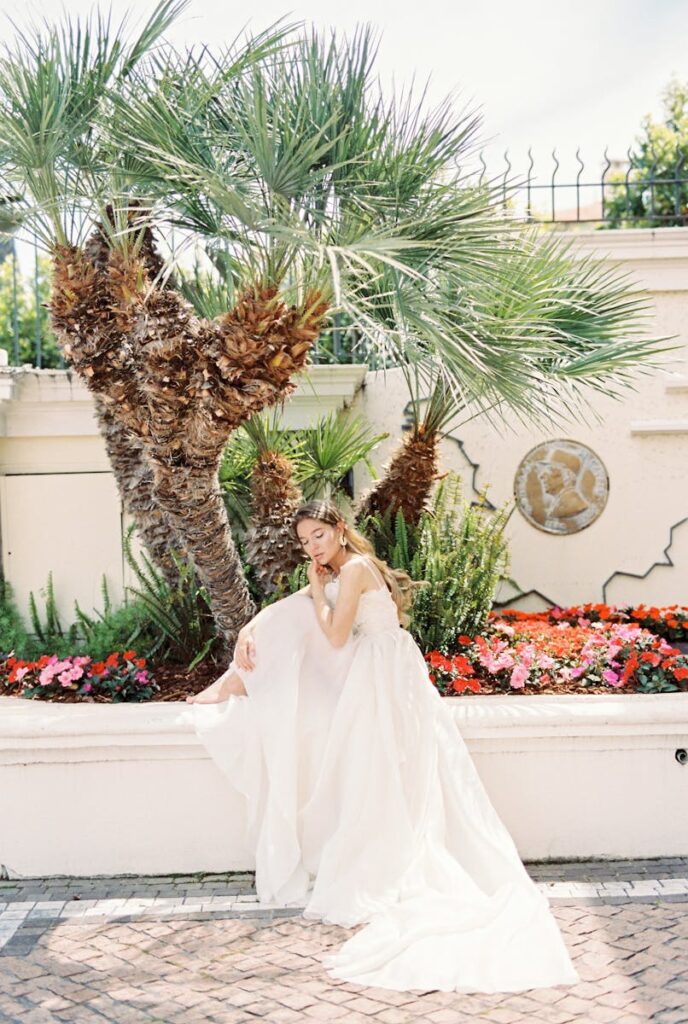 Bride elegantly seated in garden with flowers and palm tree, wearing a luxurious white wedding dress.