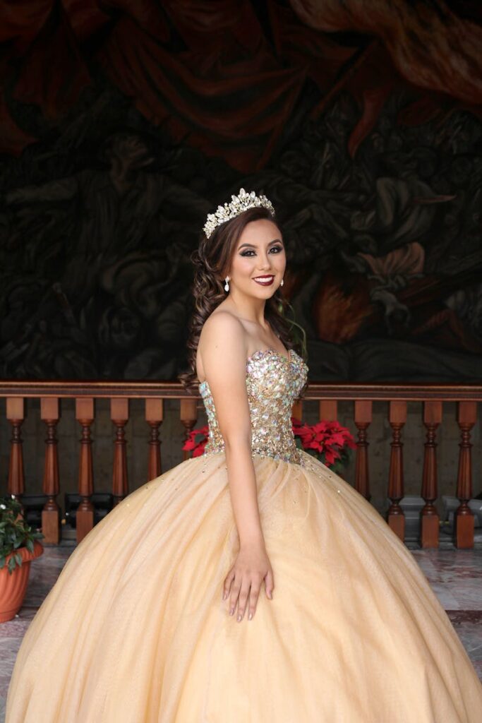 Smiling woman in a sparkling gown wearing a tiara, standing against a classic backdrop.