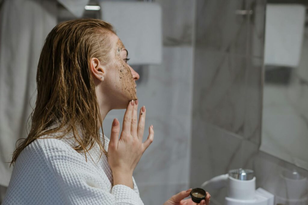 Woman with wet hair using coffee scrub on face in a modern bathroom setting.