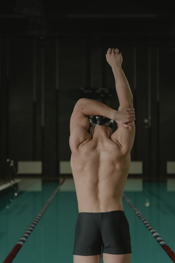 Back view of a male swimmer stretching his arm at an indoor swimming pool before training.