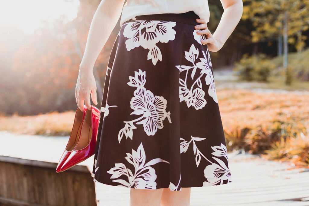 Woman holding red heels, wearing a floral skirt, enjoying a sunny day outdoors.