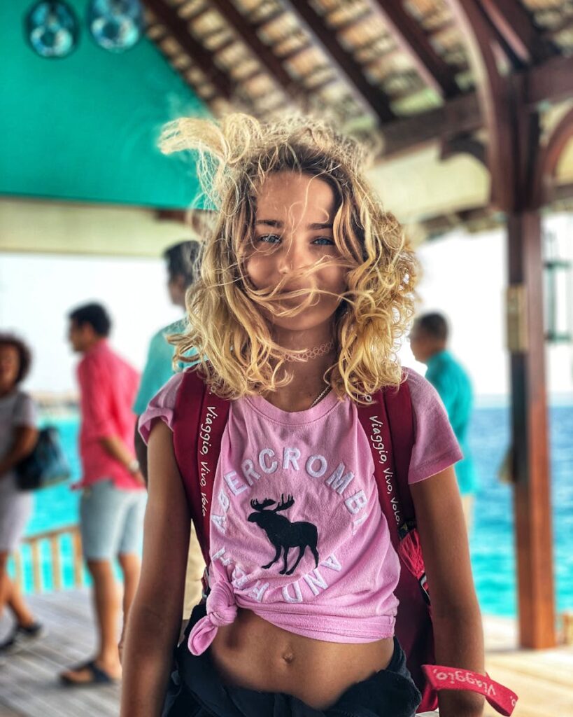 A blonde woman in the Maldives with windblown hair and a pink shirt standing by the sea.