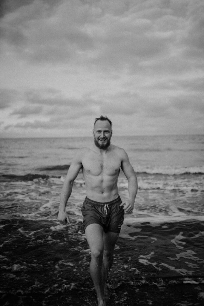 Shirtless man with beard happily walks on a beach, embracing the ocean waves under a cloudy sky.