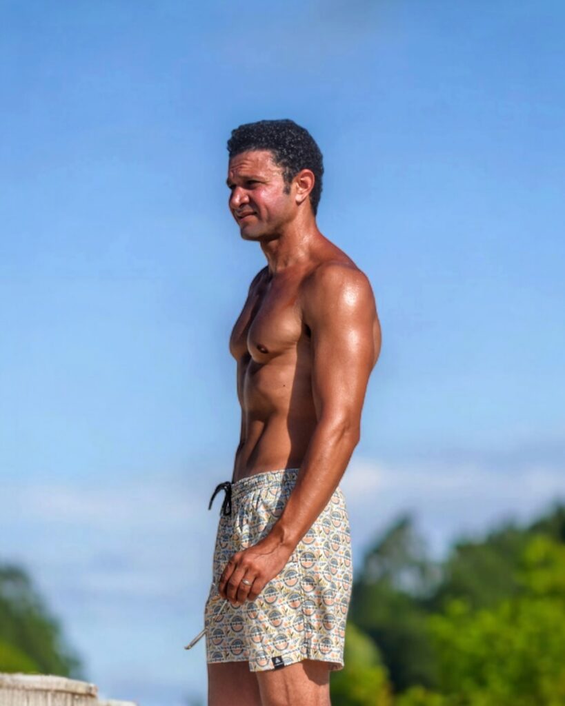 A shirtless man standing on a beach next to the ocean