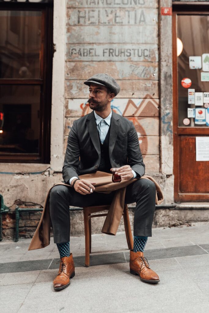 Full body of male in cap and classy suit with cigarette and tea on stool looking away on street