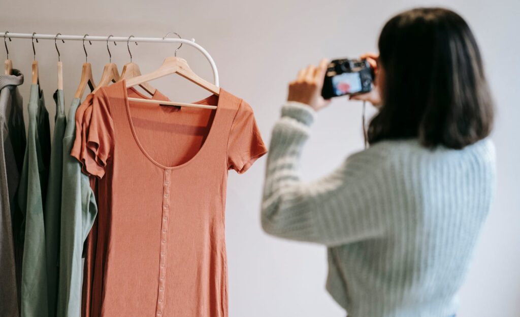 A woman photographs a clothing rack, capturing modern outfits indoors.