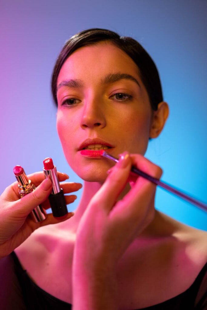 Close-up of a makeup artist applying lipstick to a woman under vibrant lighting.