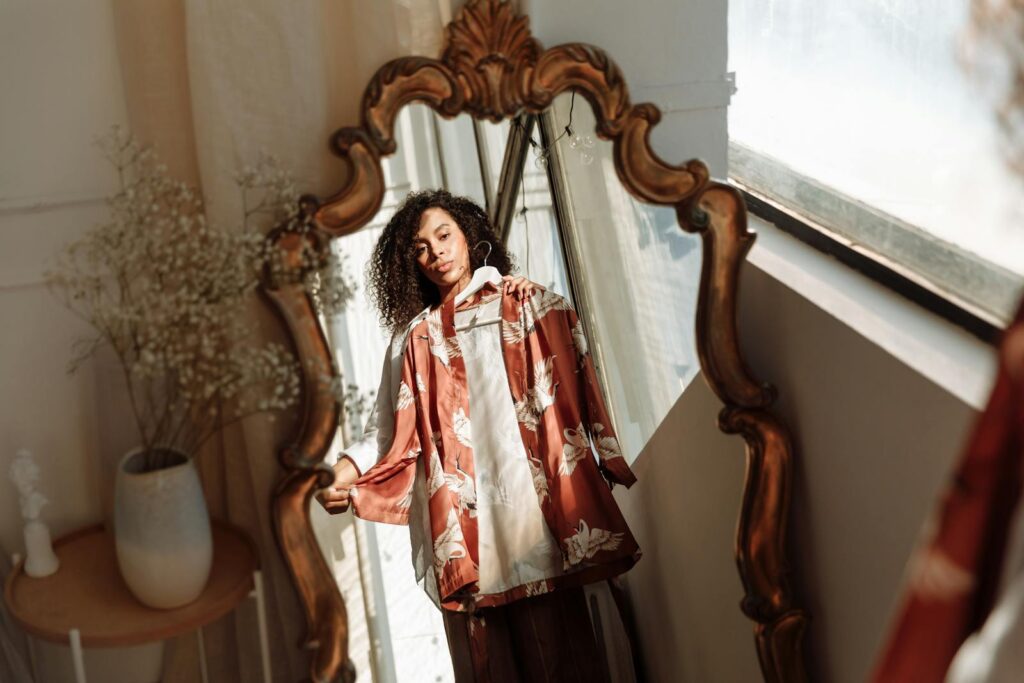 A woman holds a floral dress in front of an ornate mirror, with soft lighting enhancing the indoor setting.