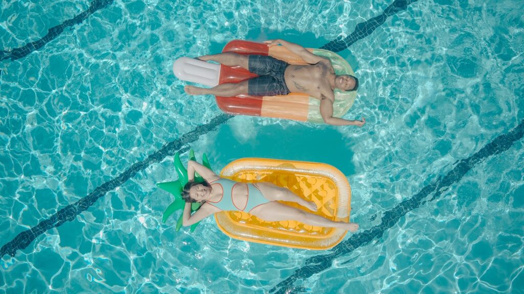Two adults relaxing on pool floats in a clear blue pool, enjoying a sunny day.