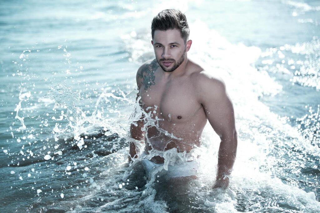 Confident young man splashes in ocean water, showing off tattoos and muscles in a dynamic beach photoshoot.