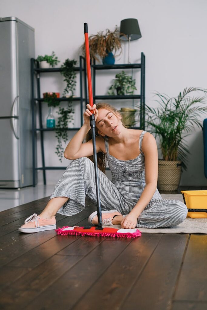 Exhausted young female with closed eyes leaning on handle of mop while sitting on carpet after washing floor in house