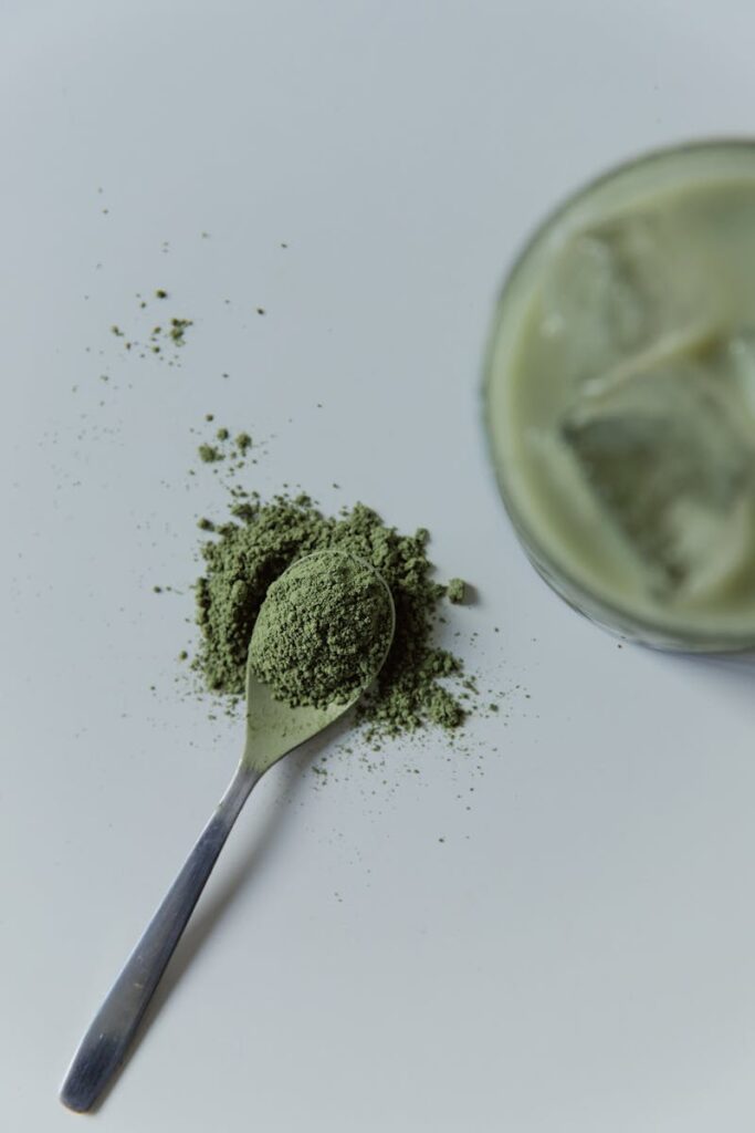 A close-up of matcha green tea powder with a spoon and iced drink on a white surface.