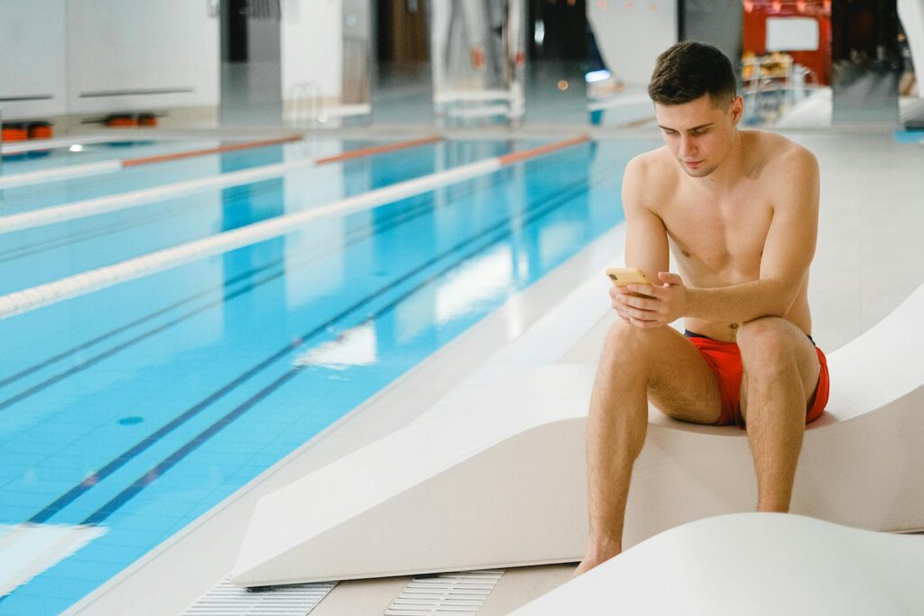 Young man in red swimming trunks using phone by an indoor pool.