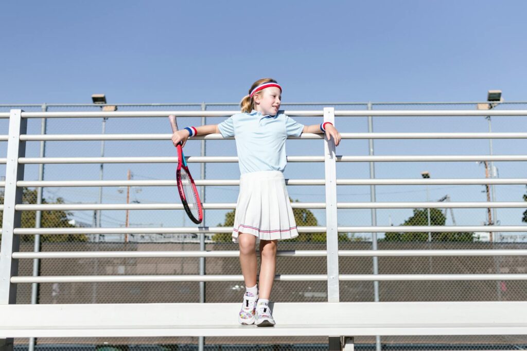 Young girl in tennis attire holding a racket, standing by a metal fence outdoors.