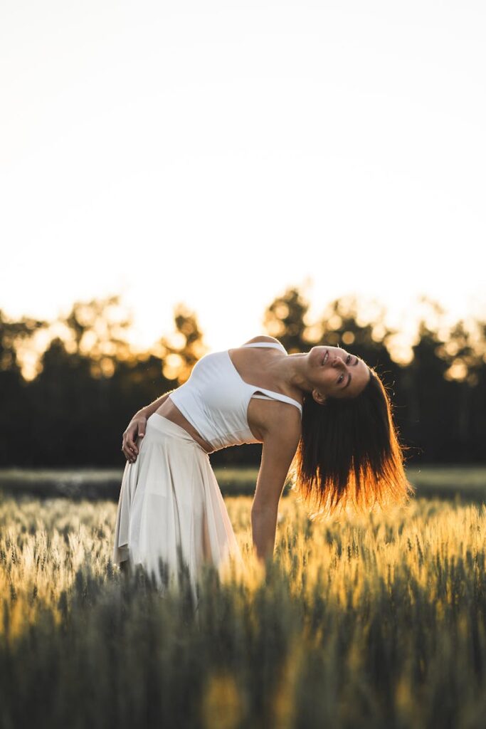 A woman with long hair in a white dress poses gracefully in a sunlit field during sunset.