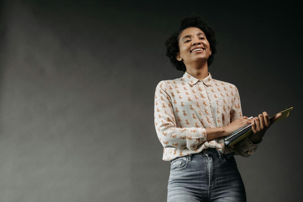 Smiling woman in a patterned blouse and jeans holding a notepad, posing confidently in a studio setting.