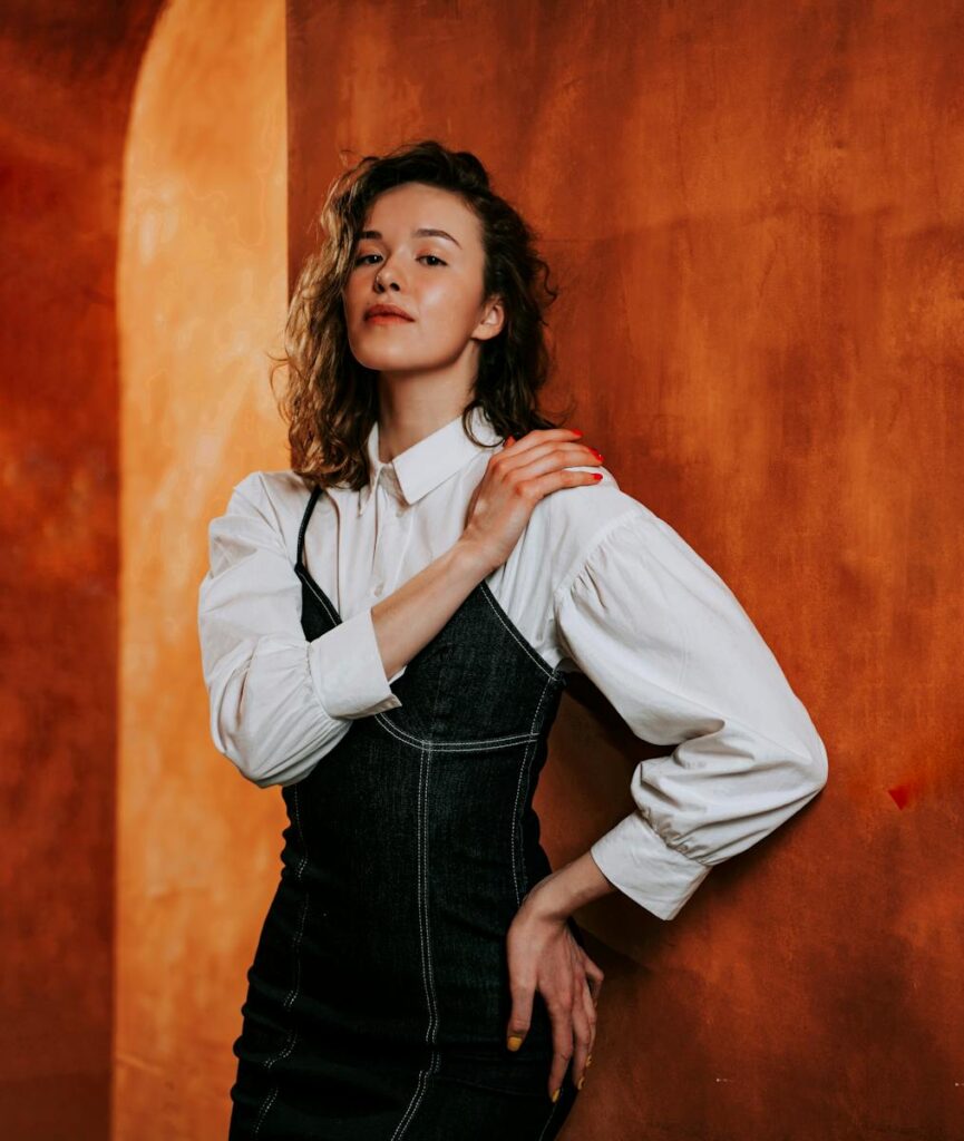 Stylish woman with curly hair posing in fashionable attire against a textured orange wall indoors.