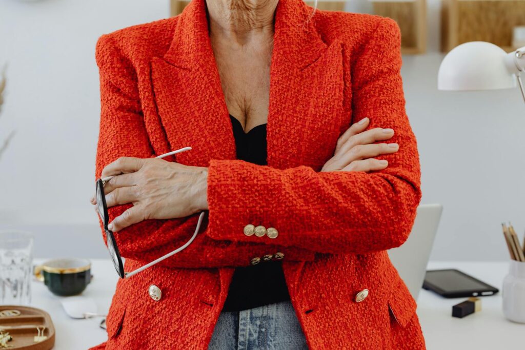 Stylish woman in bright red blazer stands confidently, holding eyeglasses indoors.