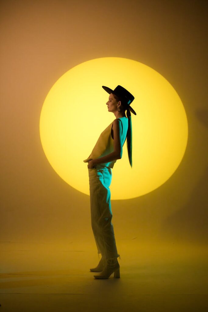 Elegant woman with hat in moody studio lighting against yellow backdrop.