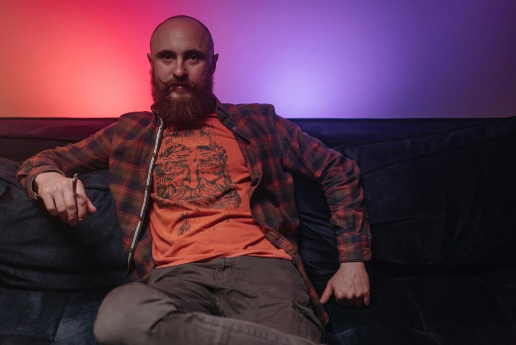 Bearded man in plaid shirt sitting on couch with colorful ambient lighting.