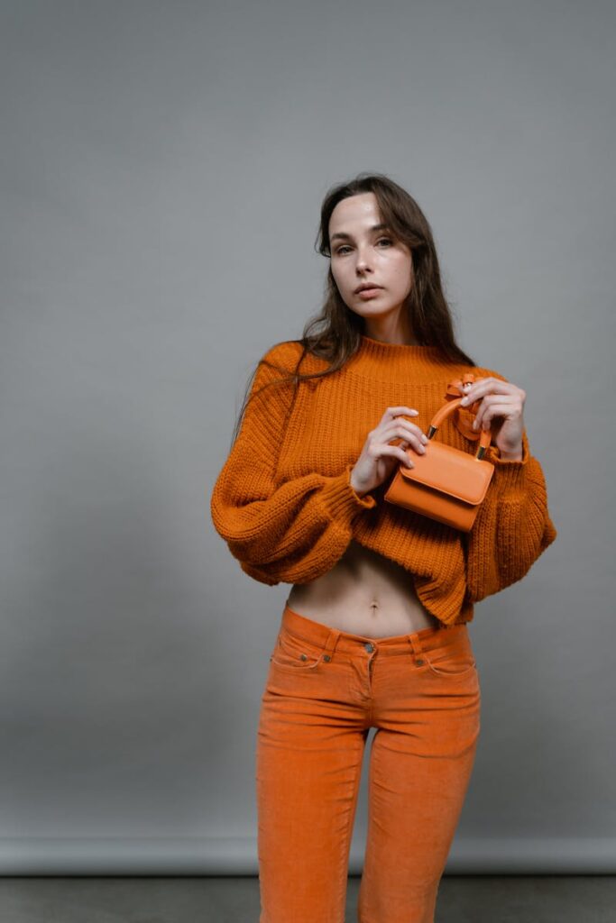 Stylish woman posing in a studio with an orange knitted sweater and micro bag, showcasing fashion trends.