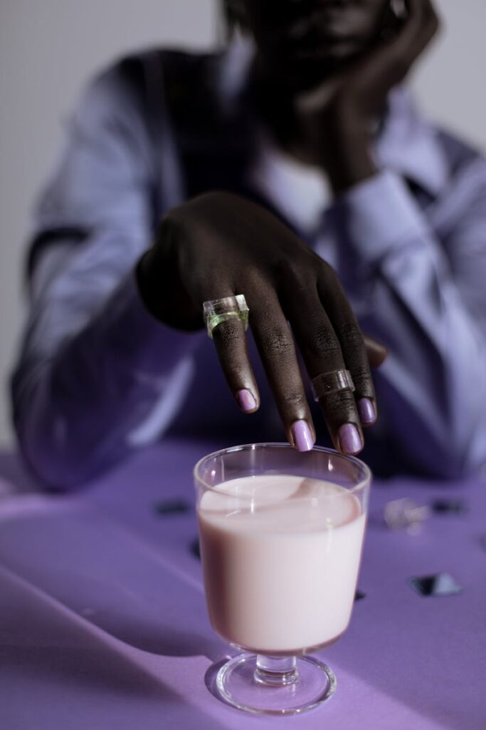 Close-up of a hand with manicured nails holding a glass of milk on a purple table.