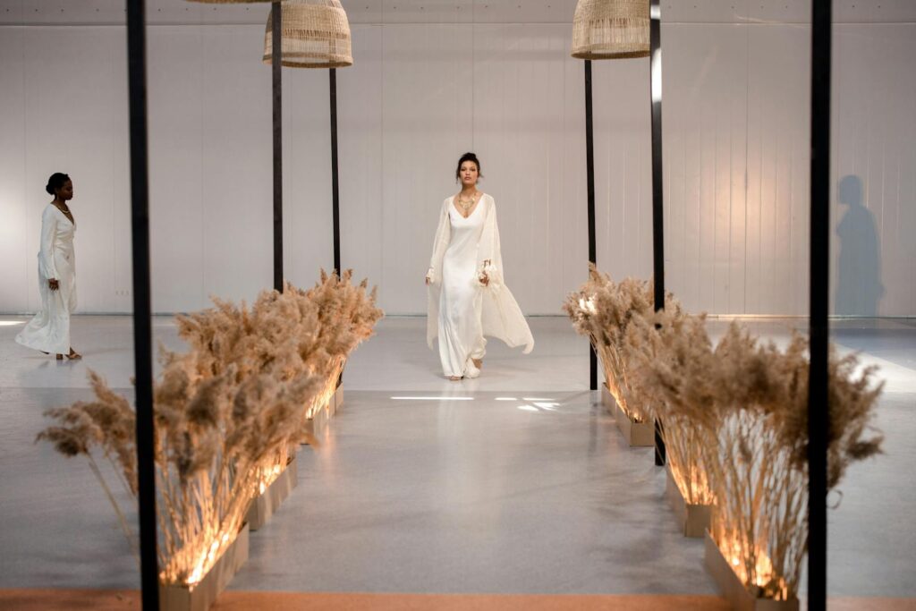 Models walking down a stylish runway in white dresses during an elegant indoor fashion show.