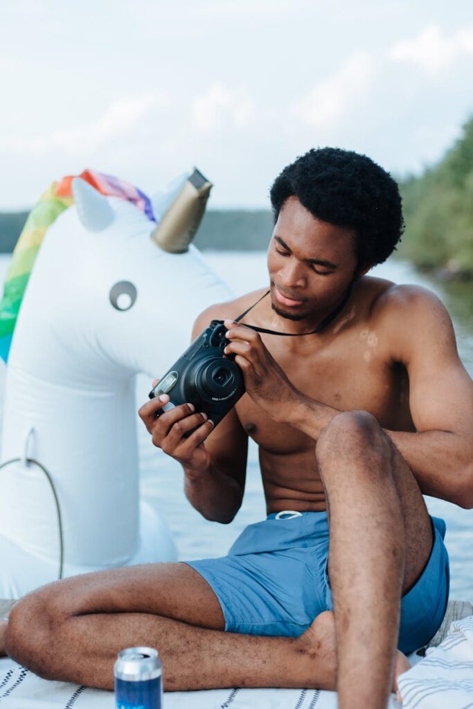 A man enjoying a sunny day by the lake, sitting with a Polaroid camera and inflatable unicorn.