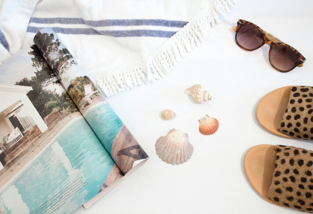 opened magazine beside seashells, slide sandals, sunglasses and towel on white surface