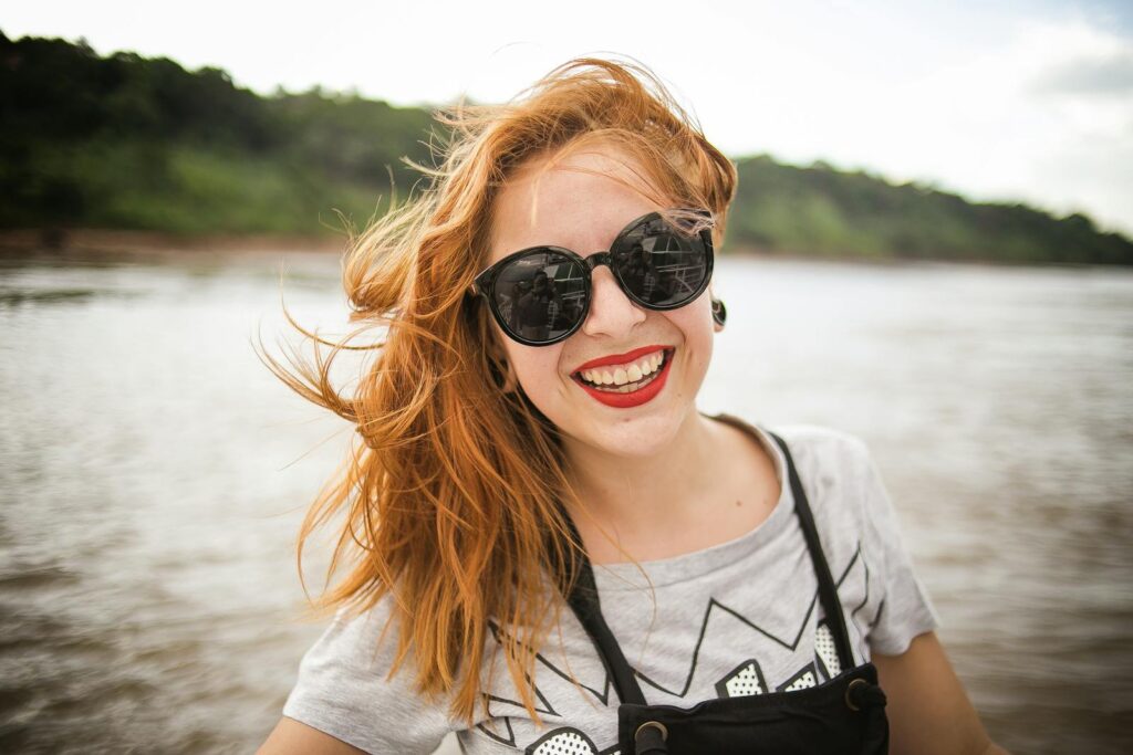 Smiling young woman with sunglasses and wind-swept hair enjoying a sunny day by the lake.