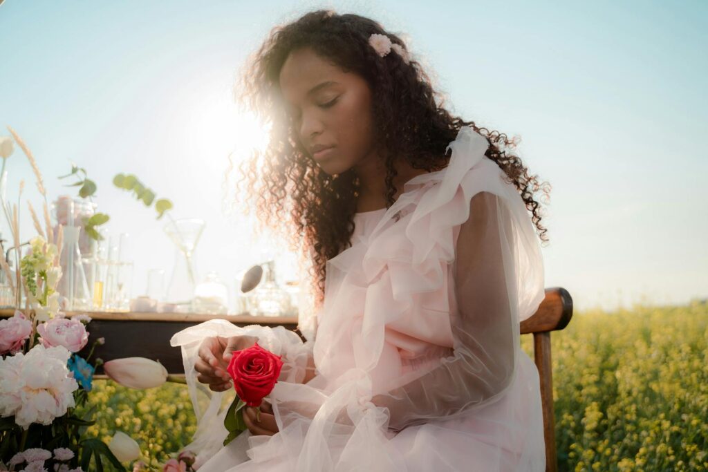 Elegant woman in a pastel dress holding a rose outdoors in a soft, dreamy light setting.