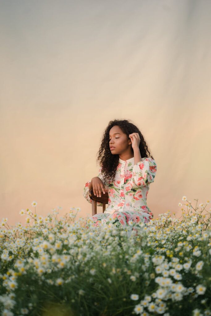 Serene woman in floral dress with curly hair sitting among daisies at sunset.
