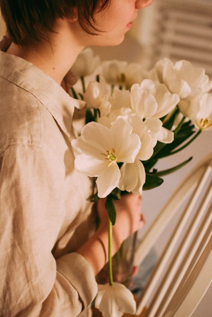 A woman in a linen shirt holds a bouquet of white flowers, symbolizing purity and elegance indoors.