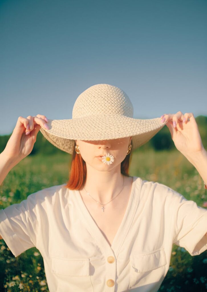 A woman enjoys a sunny day wearing a straw hat while holding a daisy flower in a vibrant meadow.