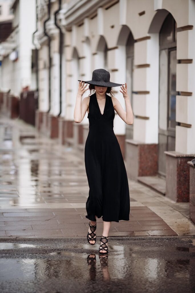 Fashionable woman in black dress and hat walking along a wet city street.