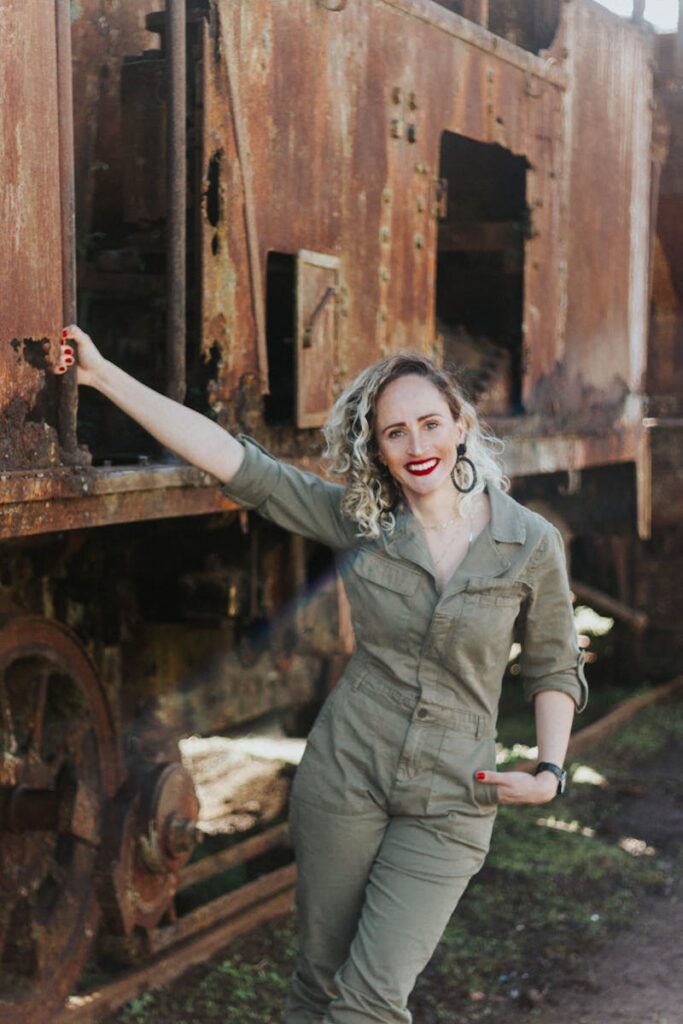 Smiling woman in green jumpsuit poses next to a rusty old train outdoors.