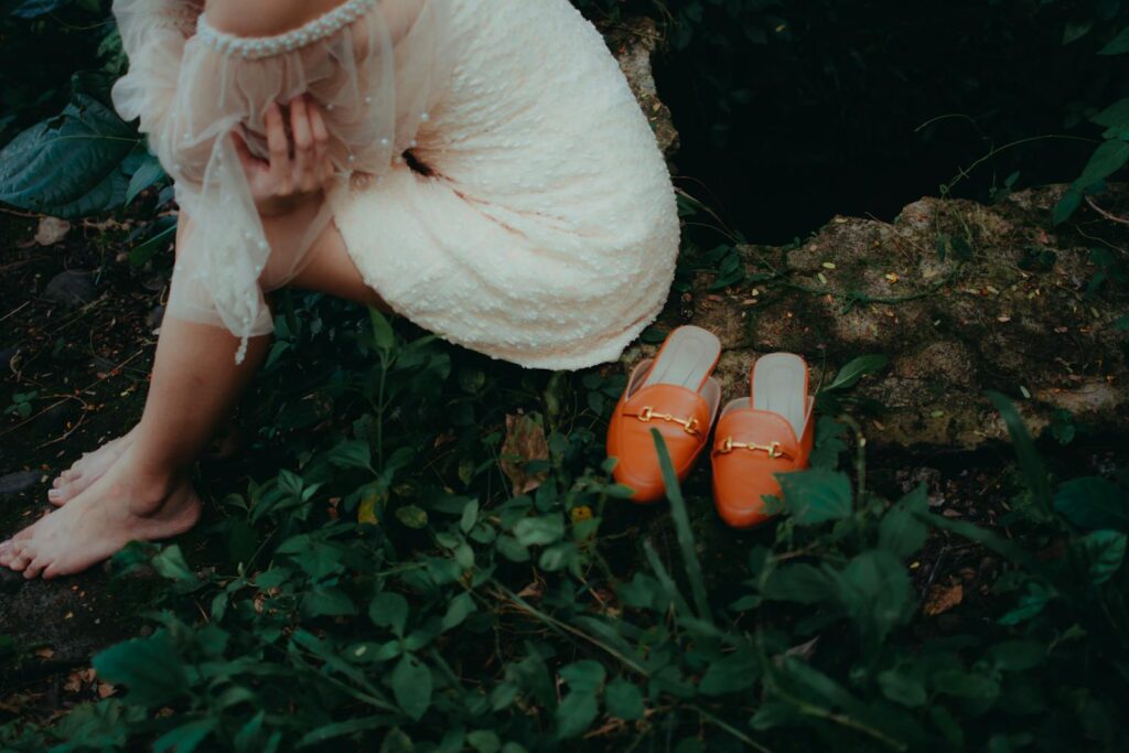 A woman in a lace dress sitting barefoot in the forest near orange loafers.