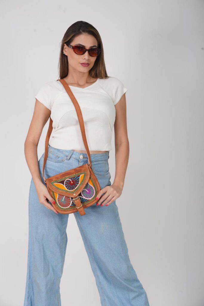 Model in sunglasses and wide jeans showcasing a vibrant handbag in a studio setting.