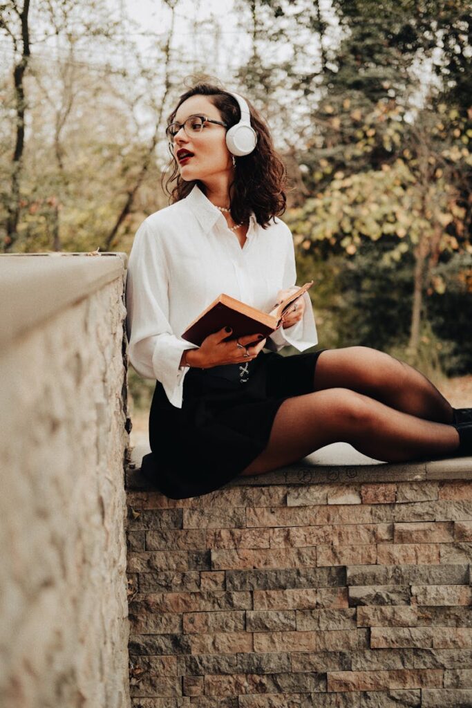 Woman in stylish outfit reading a book and listening to music outdoors.