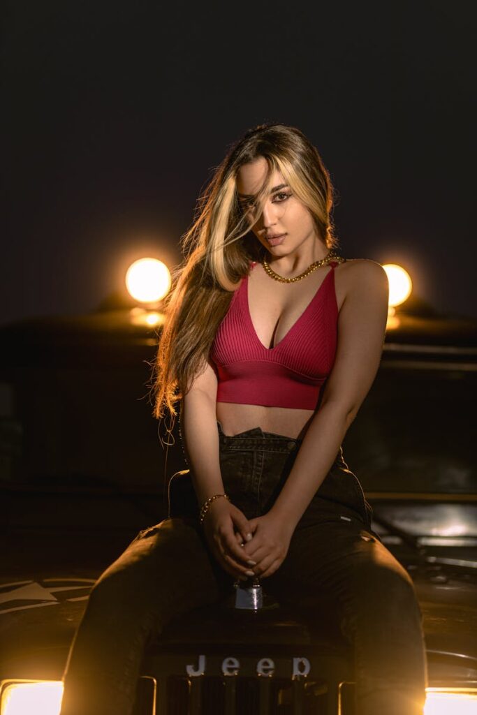 Young woman in a red crop top sitting on a Jeep at night with bright lights.