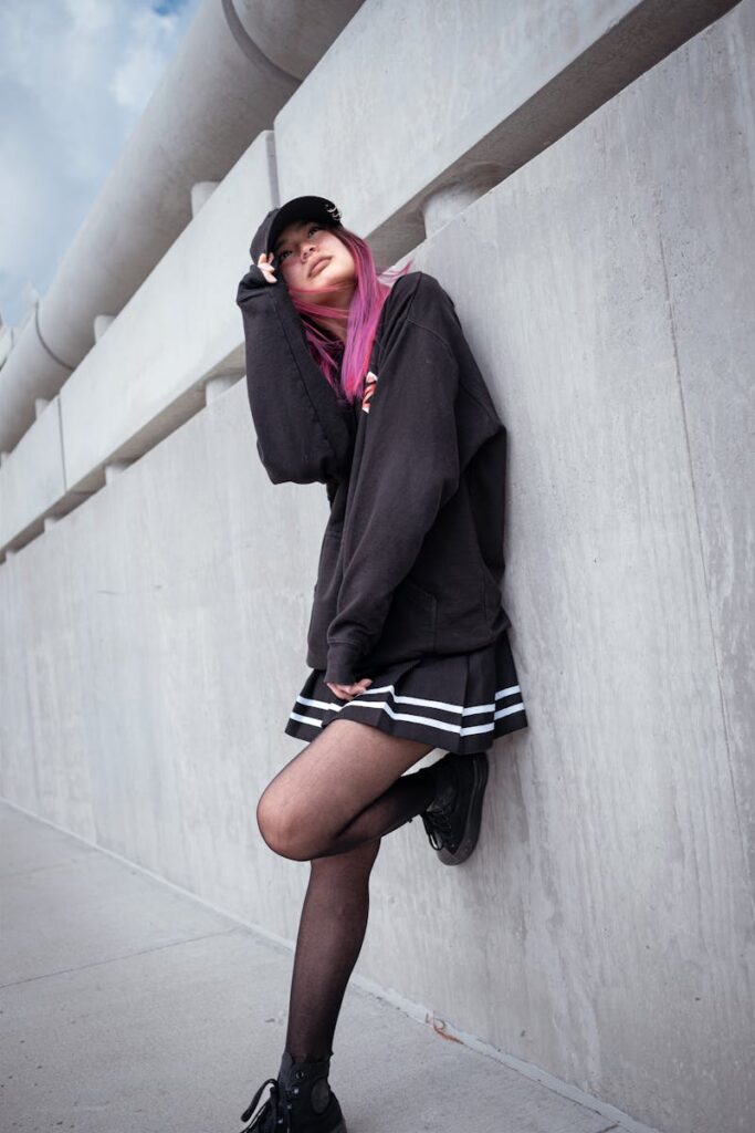 A stylish woman with pink hair leans against a concrete wall, wearing a black jacket and mini skirt.