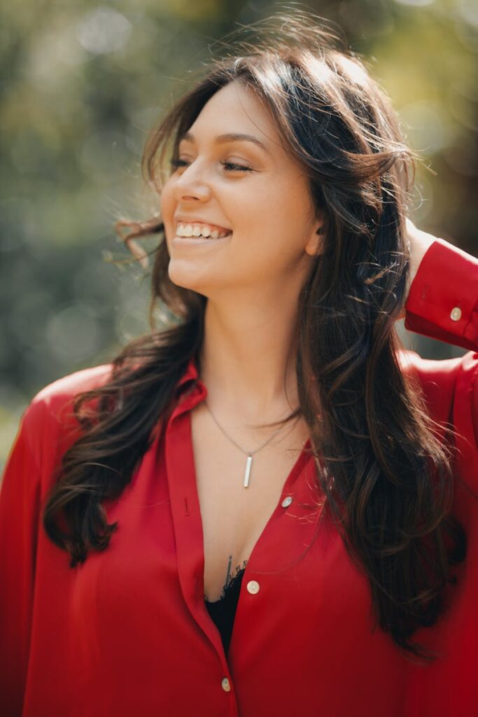 A young woman with long dark hair smiling while wearing a red blouse, captured in natural sunlight.