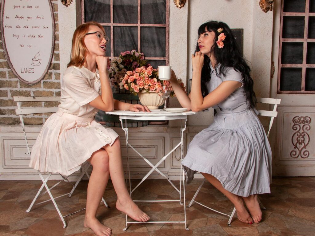 Two elegant women wearing vintage dresses enjoying conversation and coffee at a cozy indoor café setting.