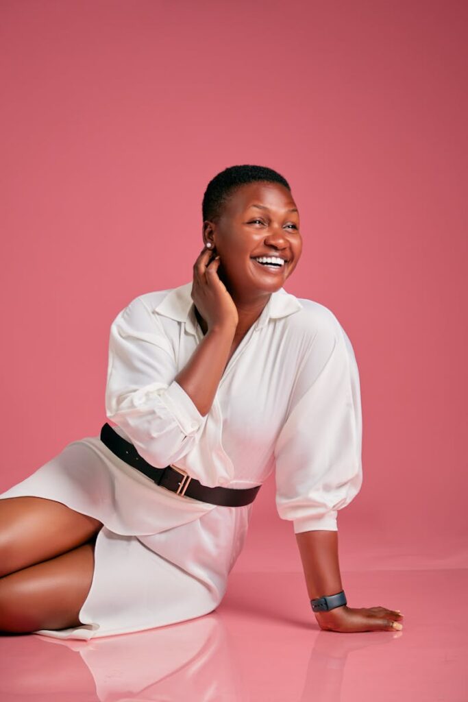 Black woman in a shirt dress sitting and laughing against pink background in a studio setting.