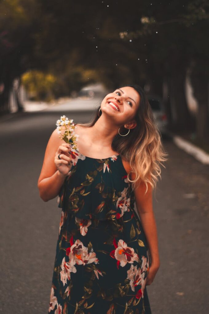 Smiling woman in floral dress holding flowers on a street. Captures happiness and summer vibes.