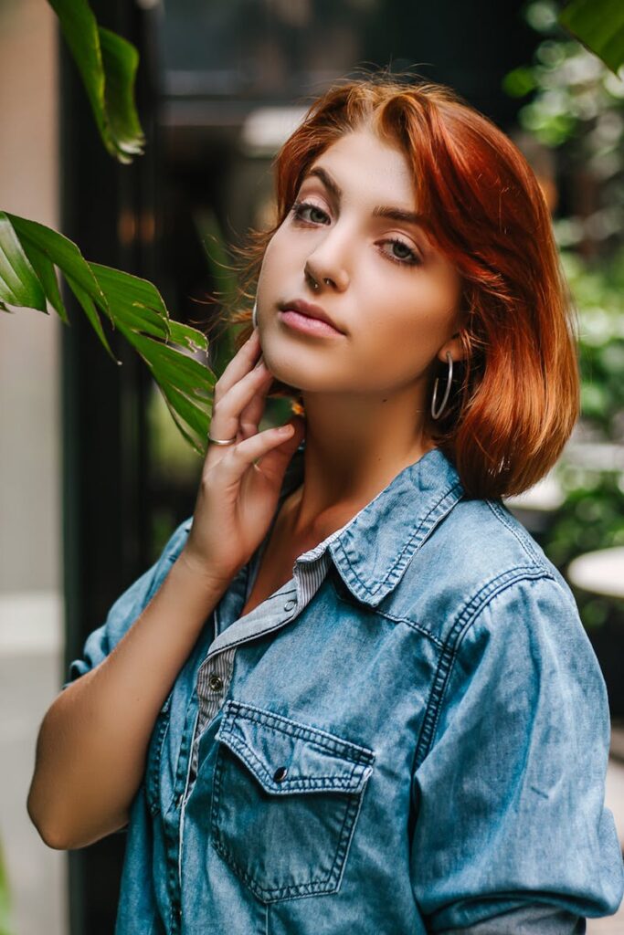 Portrait of a fashionable young woman with red hair posing outdoors in a denim shirt.