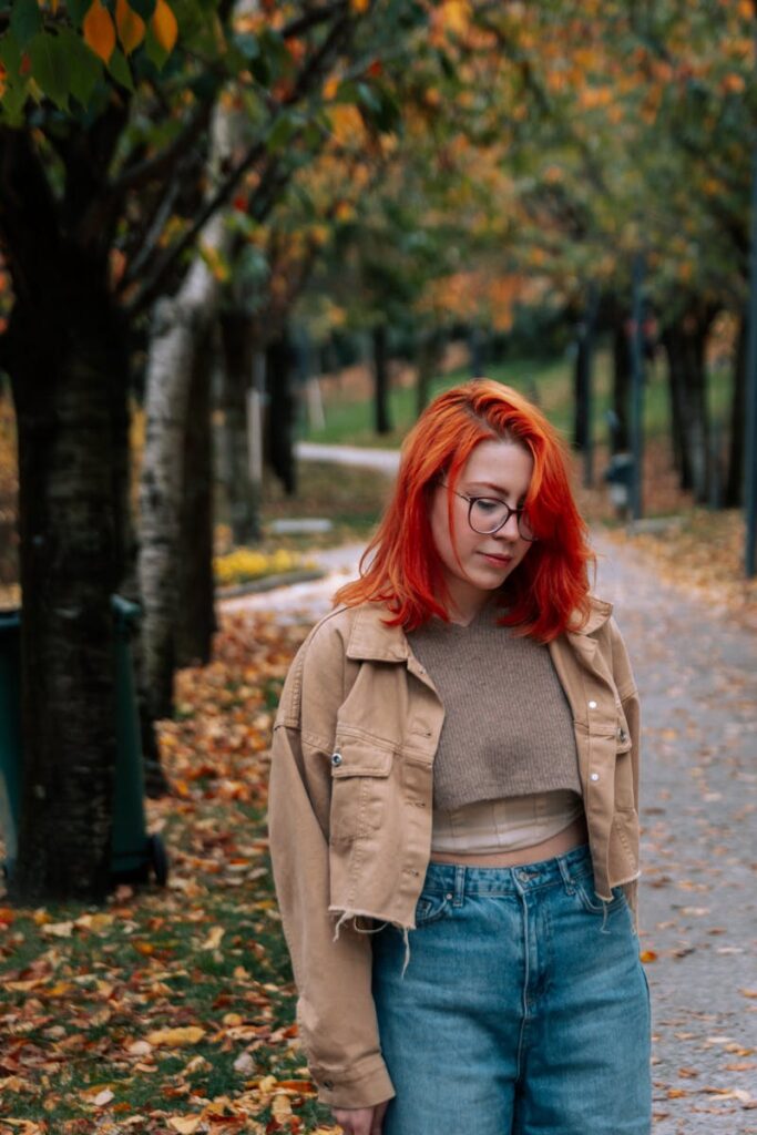 Fashionable woman with red hair in fall park, wearing a beige jacket and jeans.