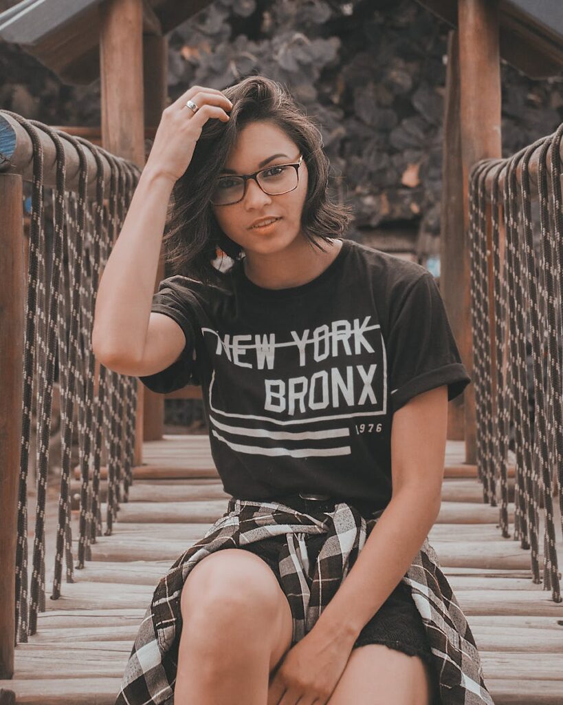 Confident woman in glasses wearing New York Bronx shirt sitting on a rustic wooden bridge outdoors.